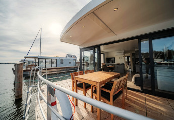 Moderne woonboot in Peenemünde met houten terras, tuinset en uitzicht op het rustige water.
