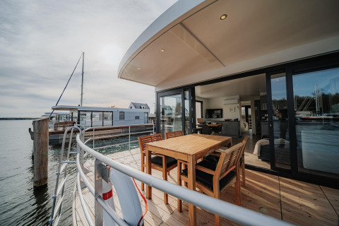Moderna casa flotante en Peenemünde con terraza de madera, comedor exterior y vistas al agua tranquila.