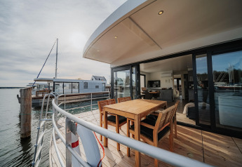 Moderna casa flotante en Peenemünde con terraza de madera, comedor exterior y vistas al agua tranquila.
