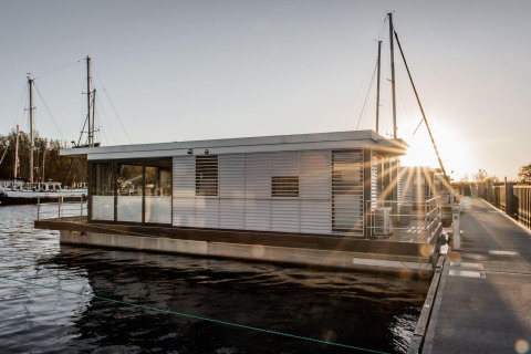 Moderne husbåd, Houseboat i Peenemünde, fortøjet ved en mole i solnedgang med sejlskibe i baggrunden.