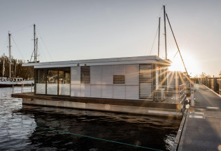 Moderne woonboot, Houseboat in Peenemünde, aangemeerd aan een dok bij zonsondergang met zeilboten op de achtergrond.