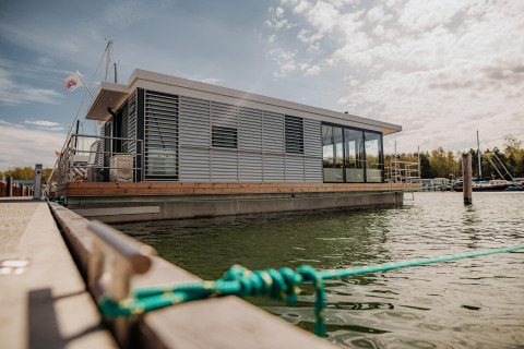 Moderne husbåd i Peenemünde fortøjet ved kajen, omgivet af vand og marina, under en let overskyet himmel.