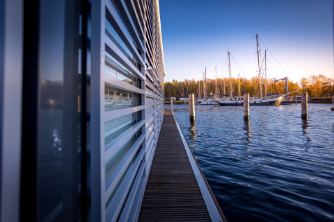 Udsigt fra husbåd i Peenemünde, viser en gangbro langs huset og både forankret i det rolige vand.