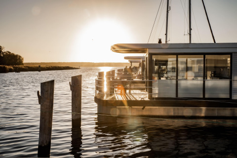 Sun setting behind a modern houseboat docked in Peenemünde, reflecting on calm water and serene scenery.