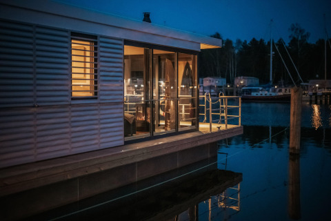 Modern houseboat in Peenemünde, warmly lit at night, with large glass windows and a serene waterfront view.