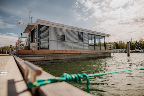 Moderne woonboot in Peenemünde, aangemeerd aan de kade, tussen het water en andere boten, onder wolken.