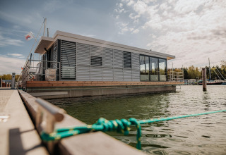 Modern houseboat in Peenemünde docked on the water, surrounded by boats and trees under a cloudy sky.