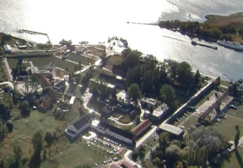 Luftfoto af en husbåd i Peenemünde med grønne områder, bygninger og sollys på vandets overflade.
