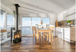 Lyst husbådinteriør med moderne køkken, spisebord, brændeovn og store vinduer med udsigt til søen.