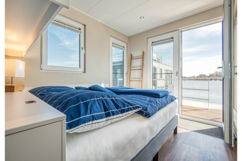 Chambre à coucher sur houseboat à Ribnitz-Damgarten, linge bleu, portes ouvertes sur vue sur l’eau.