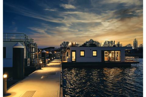 Photo of a brightly lit houseboat on the water at sunset in Ribnitz-Damgarten, with a tranquil evening sky.