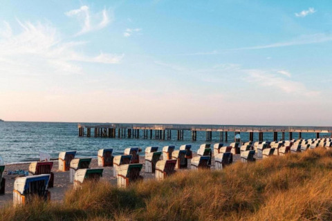 Beach at Ribnitz-Damgarten with wicker beach chairs, grassy dunes, and a pier stretching into calm sea.