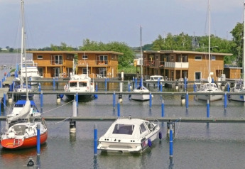 Foto de casa flotante y veleros anclados en un puerto deportivo con modernos edificios de madera al fondo.