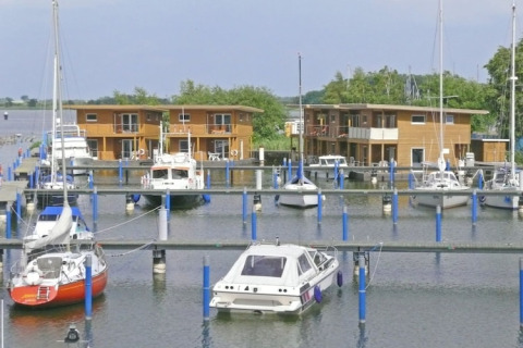 Photo d’une marina avec des péniches et voiliers amarrés, devant des maisons modernes en bois en bord d’eau.