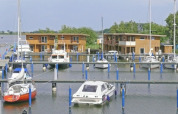 Foto van woonboten en zeilboten in een haven, met moderne houten huizen langs het water op de achtergrond.