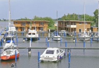 Foto von Hausbooten und Segelbooten im Yachthafen vor modernen Holzhäusern am Wasser an einem Sommertag.