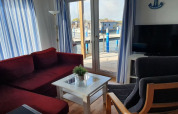 Woonkamer op een woonboot met rode bank, grijze stoel, tv, witte gordijnen en uitzicht op de haven.