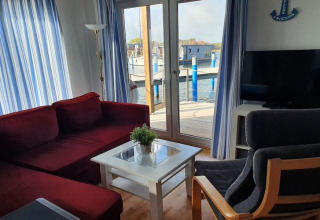 Wohnzimmer auf einem Hausboot mit rotem Sofa, grauem Stuhl, TV, weißen Vorhängen und Hafenblick.