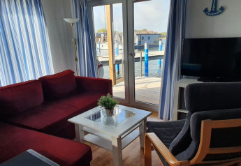 Woonkamer op een woonboot met rode bank, grijze stoel, tv, witte gordijnen en uitzicht op de haven.