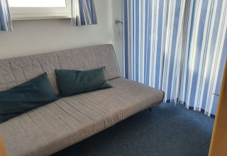 Hyggelig stue med sovesofa og blå gardiner på en husbåd. To mørke puder ligger på sofaen.