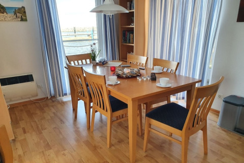 Dining area with six chairs on a houseboat, wooden floor, set table, and water view through curtains.