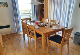 Spisebord med seks stole på en husbåd, trægulv, kopper, morgenmad og udsigt til vand gennem gardiner.