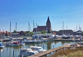 Uitzicht vanaf een woonboot op een haven met zeilboten, banken, gebouwen en een kerktoren.