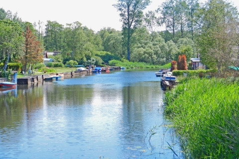 Blick auf einen ruhigen Kanal mit Hausbooten, grünen Bäumen und kleinen Booten an einem sonnigen Tag.
