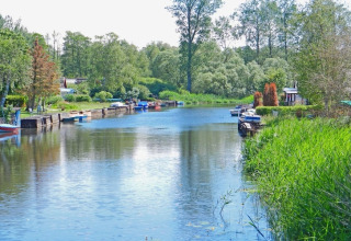 Blick auf einen ruhigen Kanal mit Hausbooten, grünen Bäumen und kleinen Booten an einem sonnigen Tag.