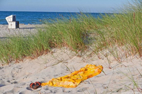 Sunny beach with dunes, grass, a yellow towel and sandals in the sand, sea and chair in background.