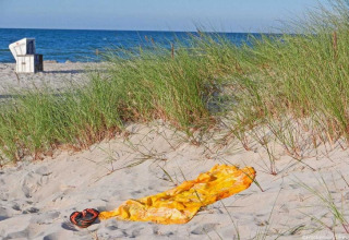 Solrig strand med klitter og græs, håndklæde og sandaler foran havet, strandstol i baggrunden.
