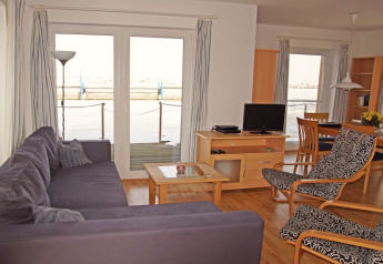 Woonkamer op een woonboot met bank, fauteuil, tv en eettafel, grote ramen met uitzicht op het water.