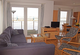 Stue på husbåd med sofa, lænestol, tv og spisestue, store vinduer med udsigt til vandet.