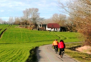 Twee fietsers op een pad langs een groen veld met een wit huis met rood dak, omgeven door bomen in Duitsland.
