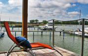 A relaxing lounge chair on a houseboat deck overlooks a marina with boats, under a partly cloudy sky.