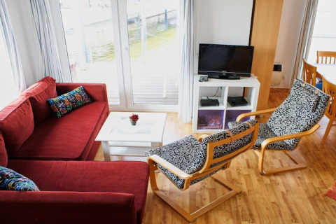 Stue på en husbåd med rødt sofaarrangement, to mønstrede stole, bord, TV og udsigt til vandet.