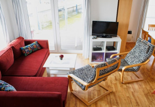Soggiorno su una houseboat con divano rosso, sedie fantasia, TV, tavolino bianco e vista sull’acqua.