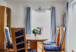 Eetruimte op een woonboot met ronde tafel, blauwe stoelen en zachte lichtinval door de gordijnen.