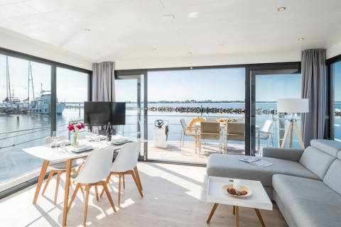 Lyst og moderne husbådinteriør med store vinduer og udsigt til havnen, spisebord og loungeområde.