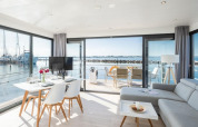 Lyst og moderne husbådinteriør med store vinduer og udsigt til havnen, spisebord og loungeområde.