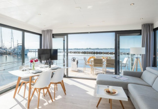Intérieur moderne de péniche avec grandes baies vitrées, coin repas, salon et vue sur le port de plaisance.