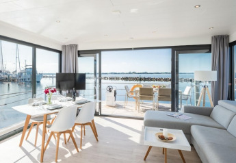 Licht en modern interieur van een woonboot met grote ramen, eettafel en zicht op de jachthaven buiten.