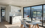 Modern woonbootinterieur met eettafel, keukenblok en grote ramen met uitzicht op de haven en boten.
