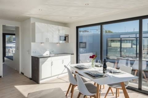 Interno di moderna houseboat con tavolo da pranzo, cucina e grandi finestre affacciate sulla marina.