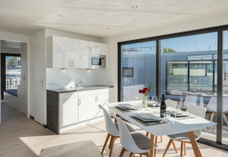 Intérieur d’une péniche moderne avec table à manger, coin cuisine et grandes fenêtres donnant sur la marina.