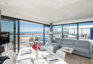 Lyst og moderne opholdsområde på en husbåd med panoramaudsigt over vandet og terrasse i solskin.