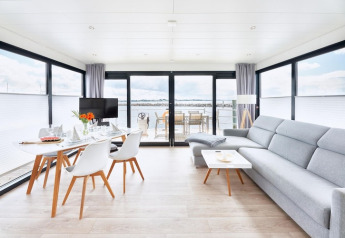 Lyst og moderne interiør på en husbåd med store vinduer, spisebord og sofa, udsigt over vandet.