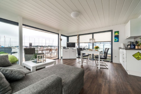 Moderne stue og køkken på en husbåd med store vinduer og udsigt over en marina med sejlbåde.