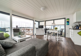 Moderne stue og køkken på en husbåd med store vinduer og udsigt over en marina med sejlbåde.