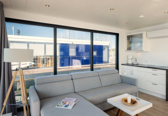 Moderne woonkamer op een woonboot met grijze bank, kitchenette en grote ramen met uitzicht op de kade.